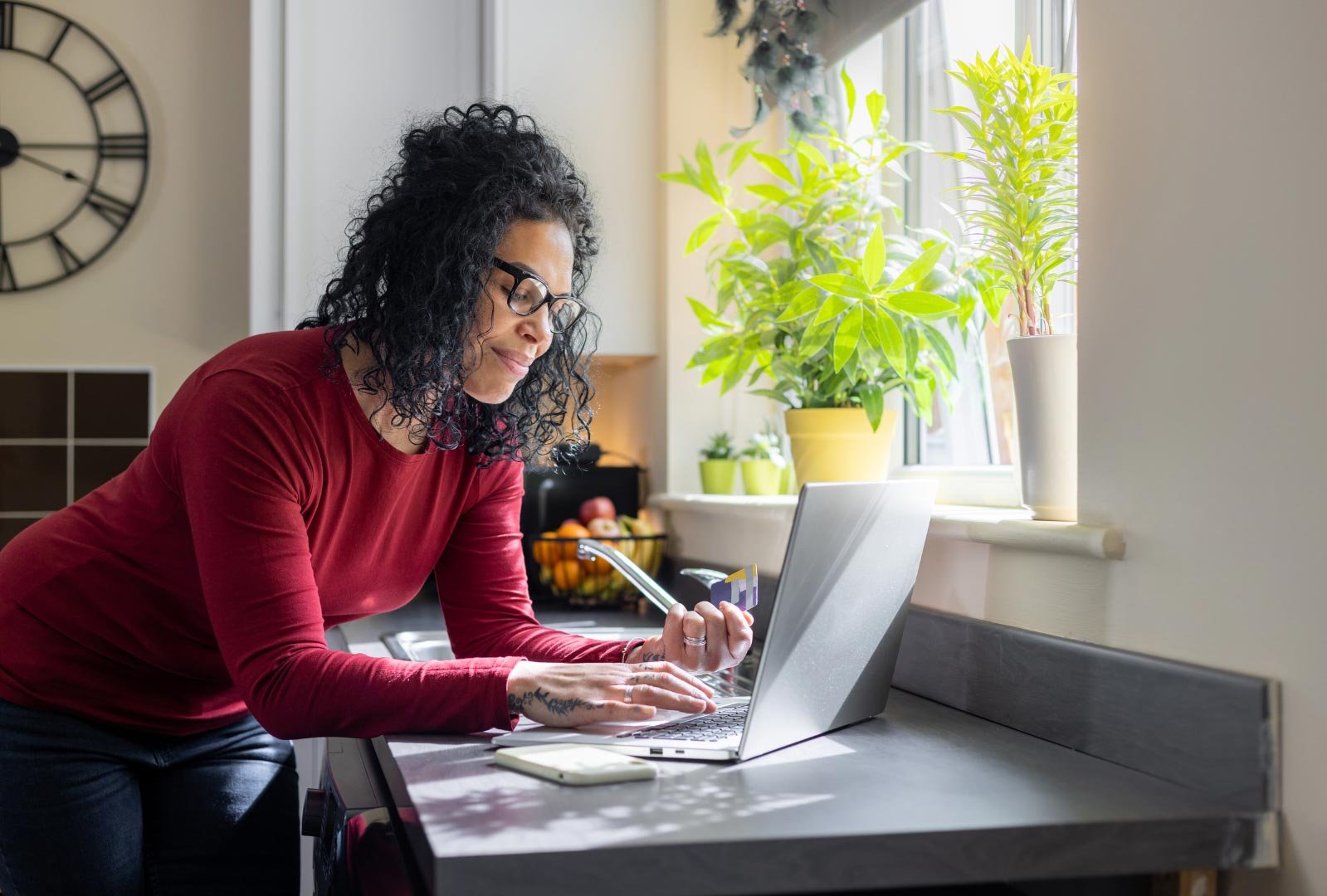 A woman working from home on her laptop in a brightly lit kitchen with plants and a clock in the background