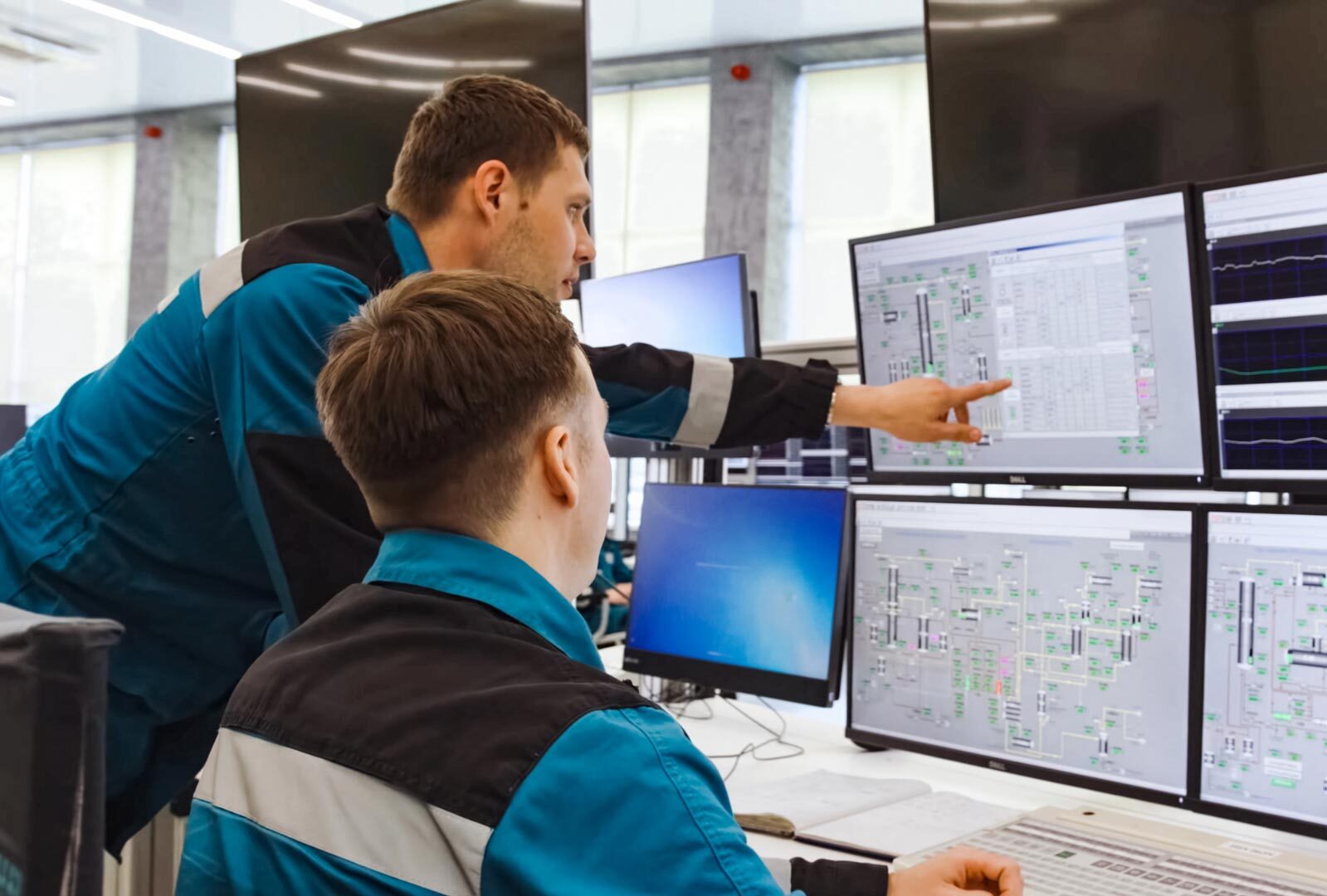 Two technicians in teal and black uniforms examining and discussing data on multiple computer screens in a control room.