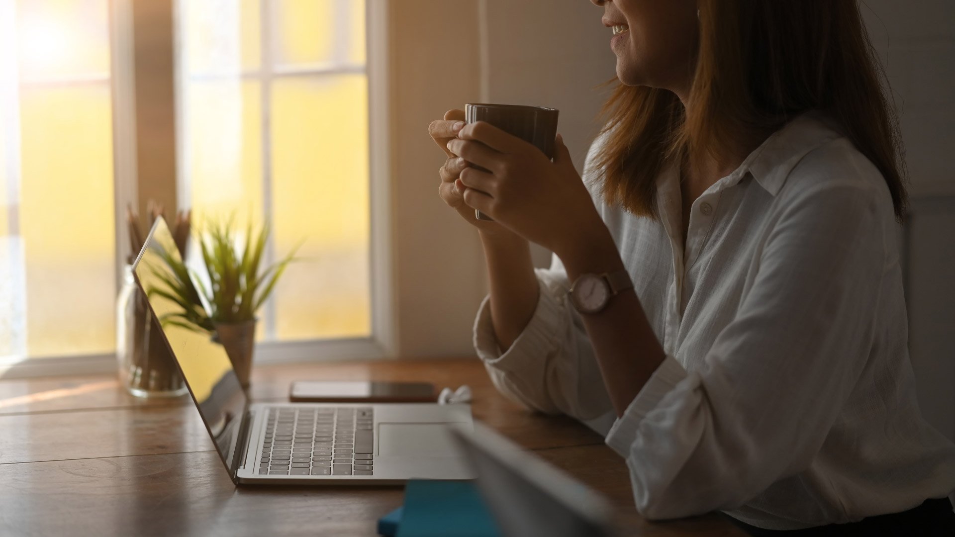 A woman enjoying her morning coffee while working on her laptop, bathed in warm sunlight.