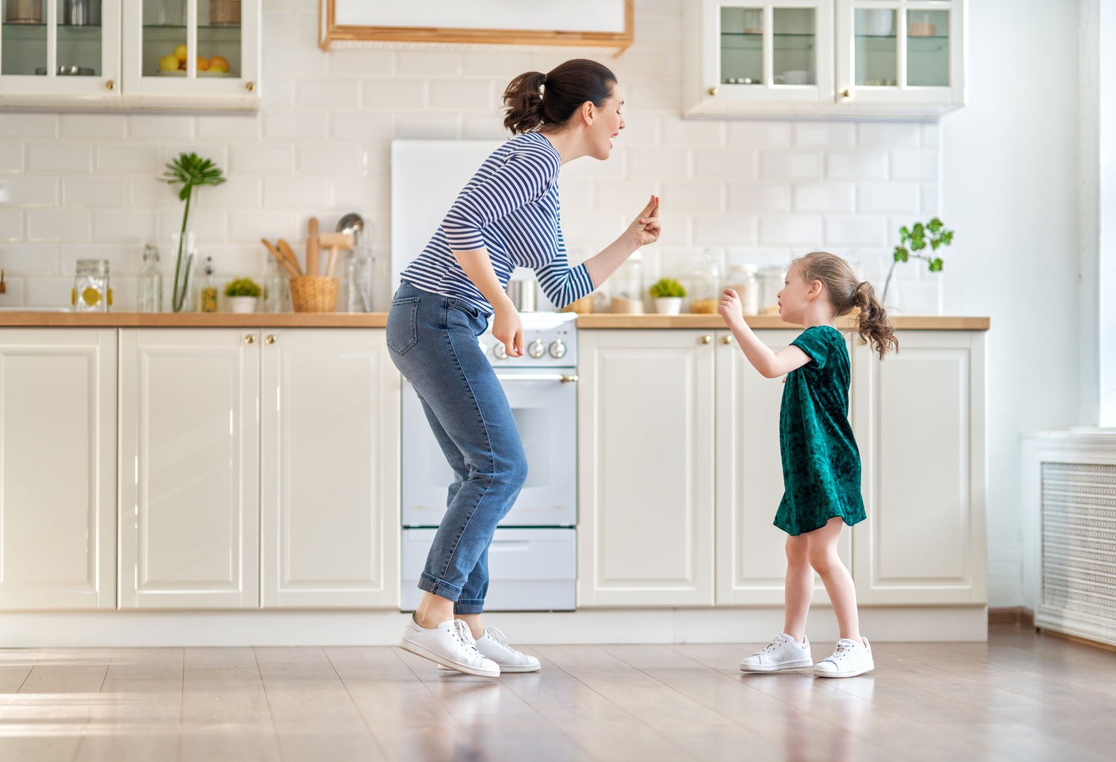 A mother and young daughter dancing happily in a bright, modern kitchen.