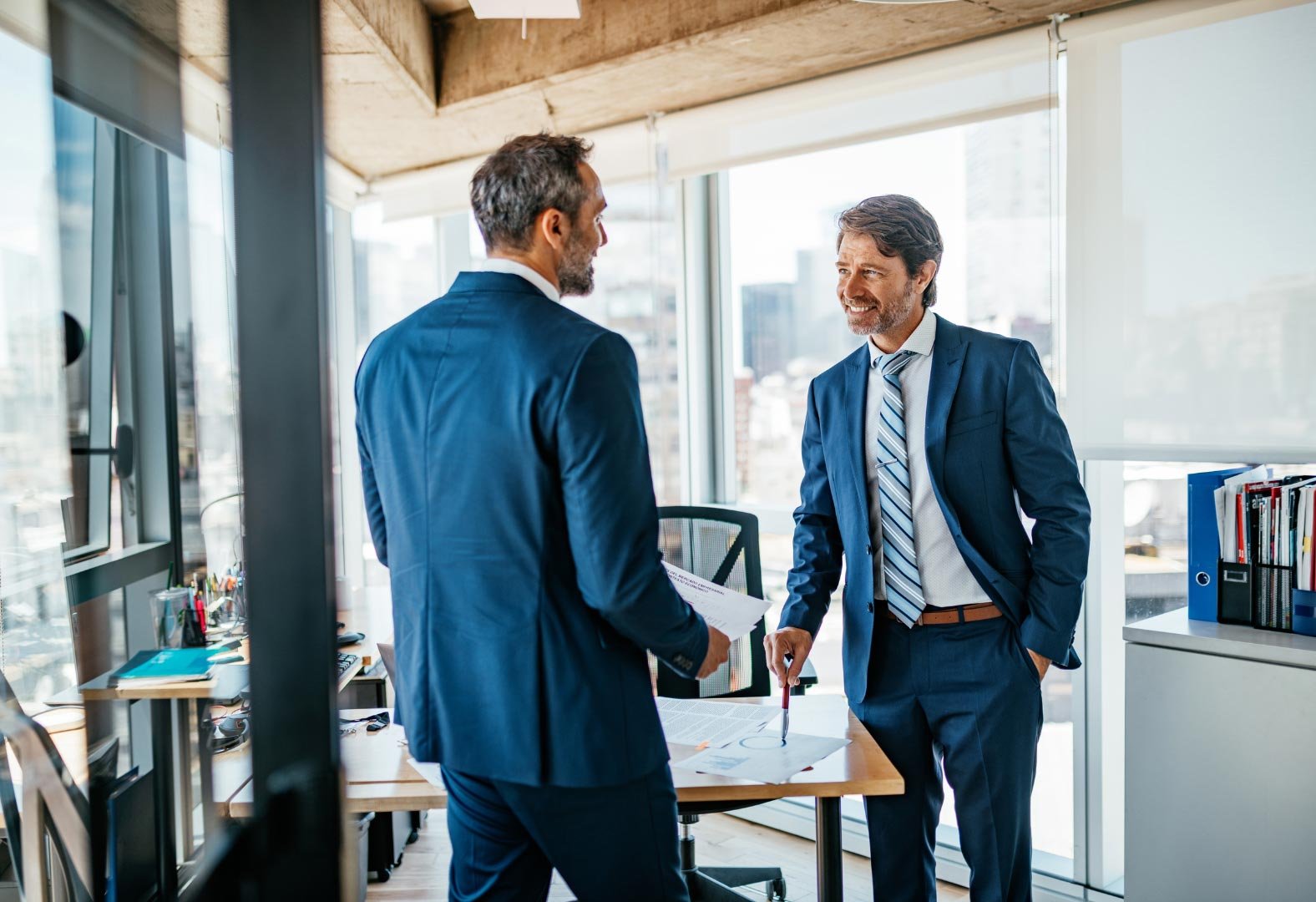 Two businessmen discussing documents in a modern office with city views