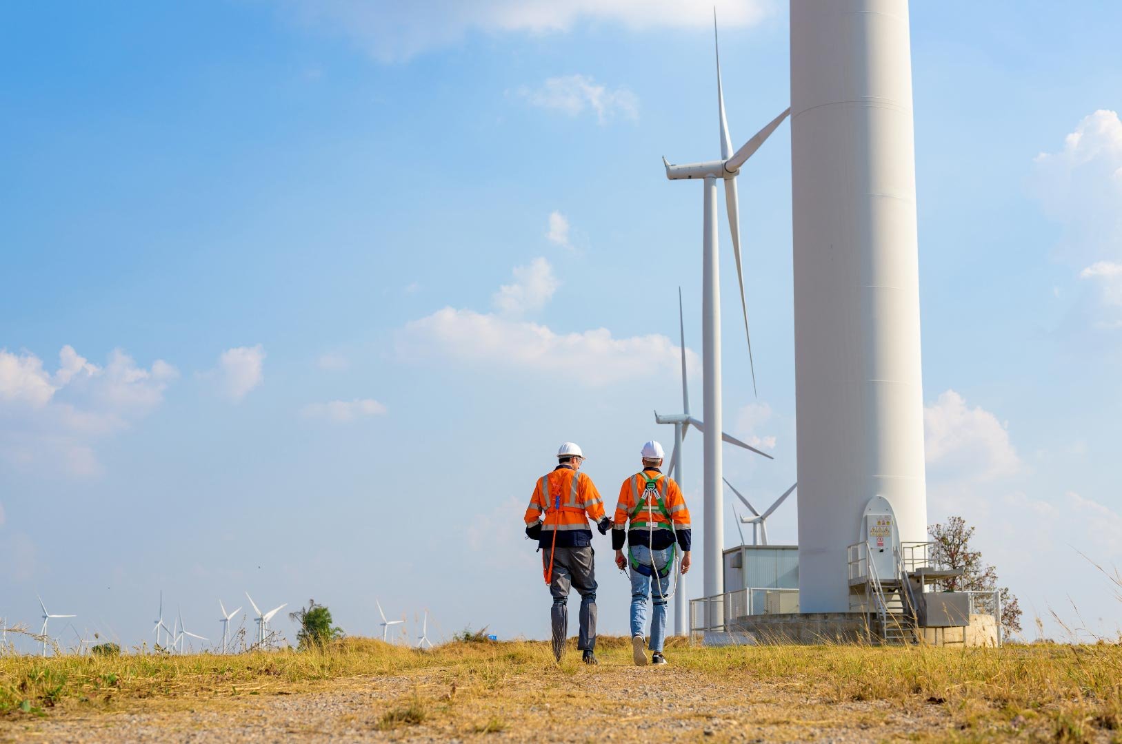 Two technicians in safety gear walking towards a large wind turbine
