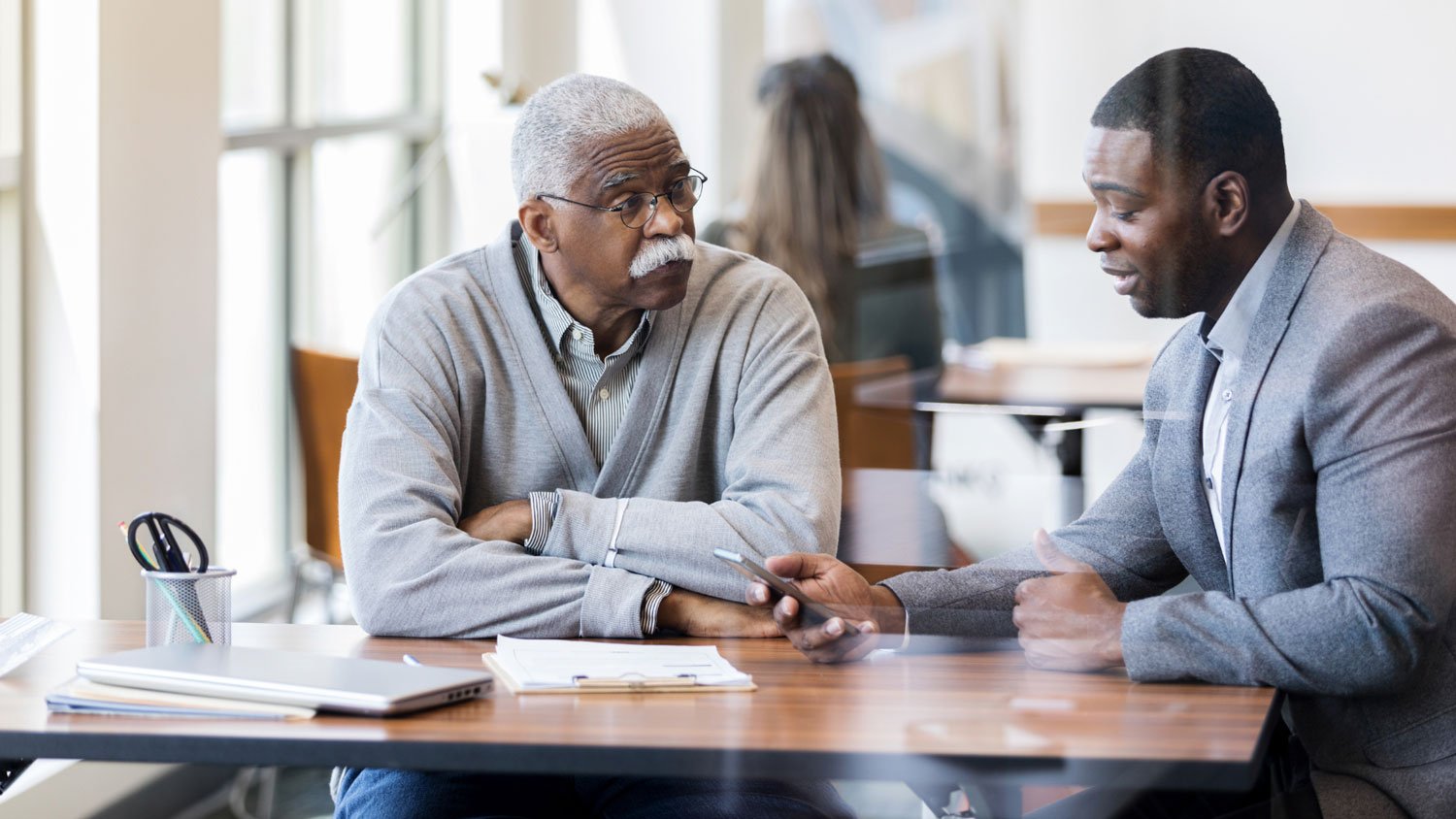 An older man and a younger man having a serious discussion at a table in a public setting.