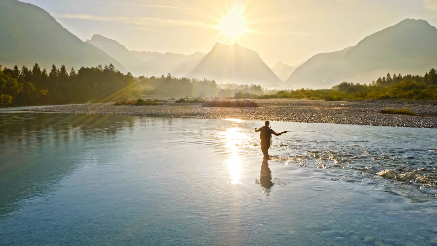 A person wading in a river casting a fishing line at sunrise with mountains in the background