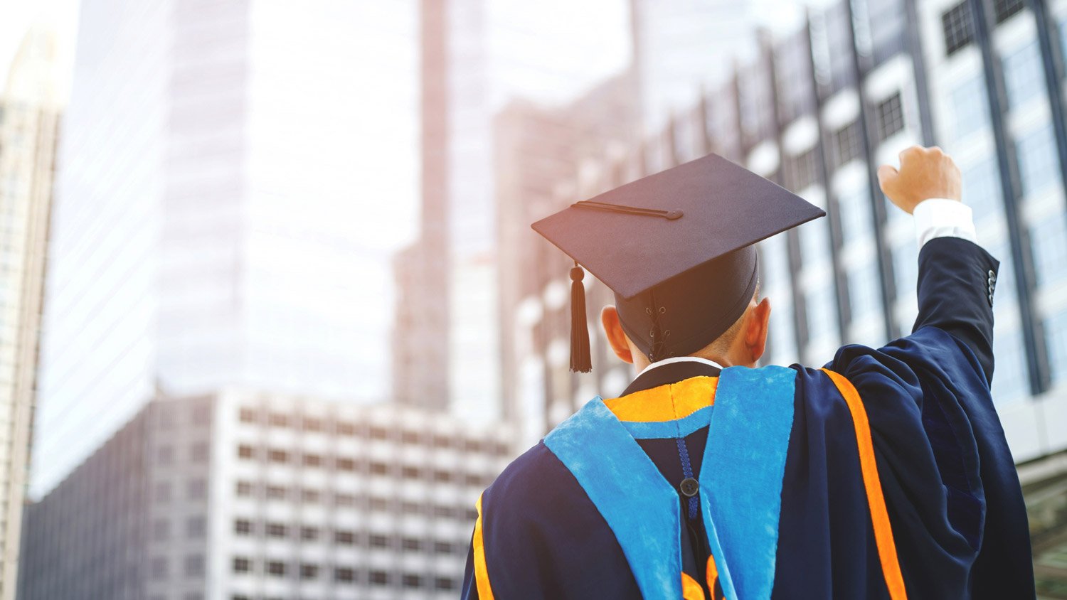 A male graduate in blue and orange regalia, fist-pumping in front of modern skyscrapers.