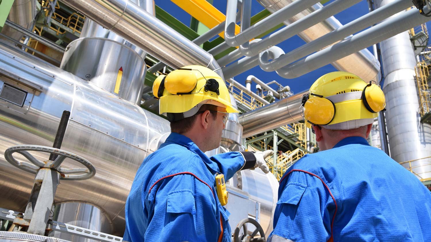Two engineers in hard hats and blue coveralls inspecting machinery at an industrial plant.