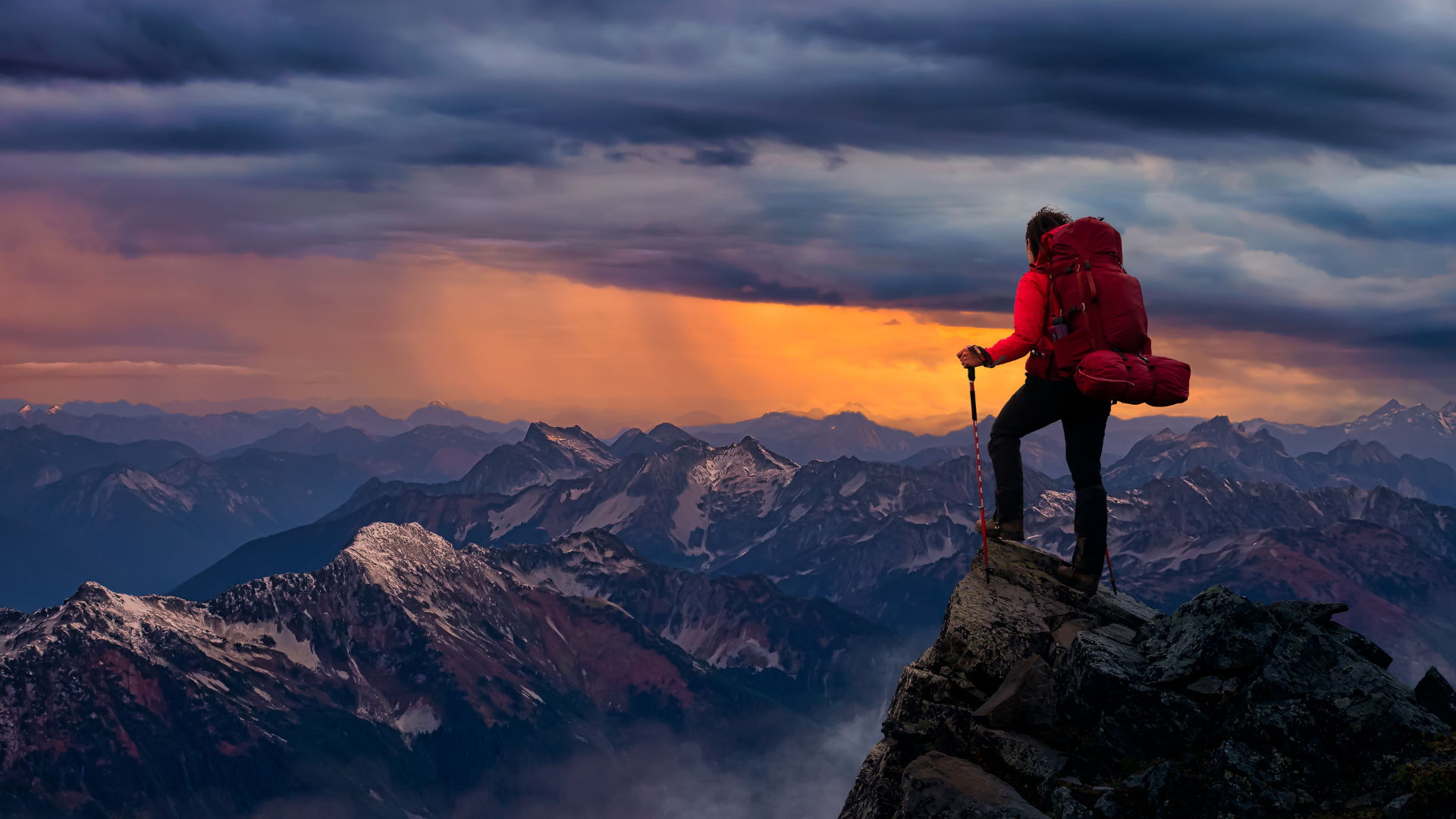 Figure with red coat and walking stick stands on peak and looks out at mountain range and sunset cloudy sky