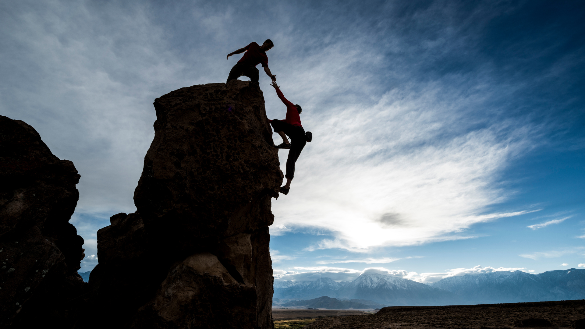 Figure reaches down to help pull climber up on top of rock pillar. Blue sky with thin wispy clouds in the background