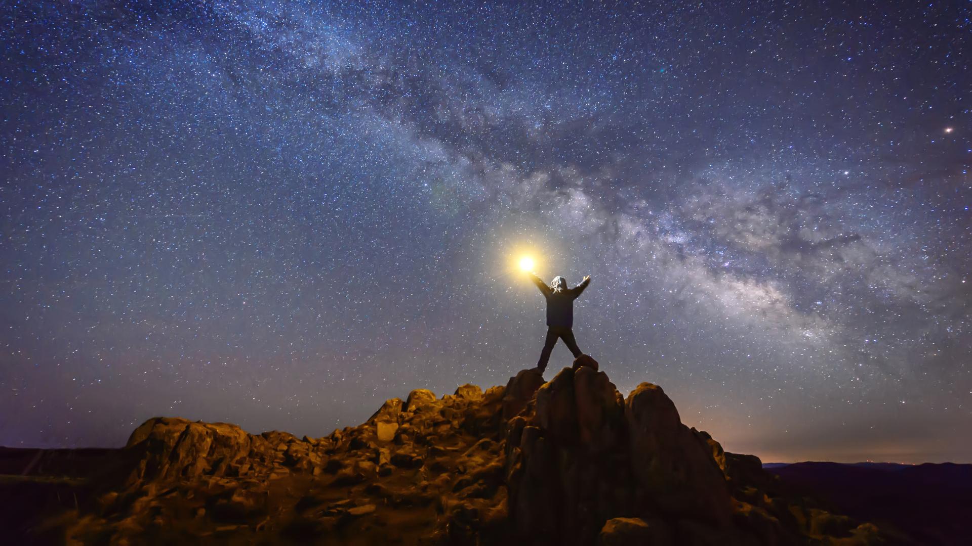 Person with light in hand standing in X shape on top of rocks, celebrating, silhouetted against a sky filled with galaxy