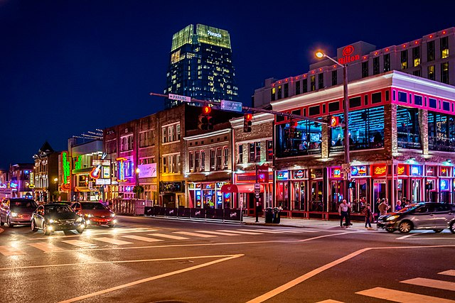 A busy city street at night with bright neon lights on buildings, cars on the road, and pedestrians on sidewalks.