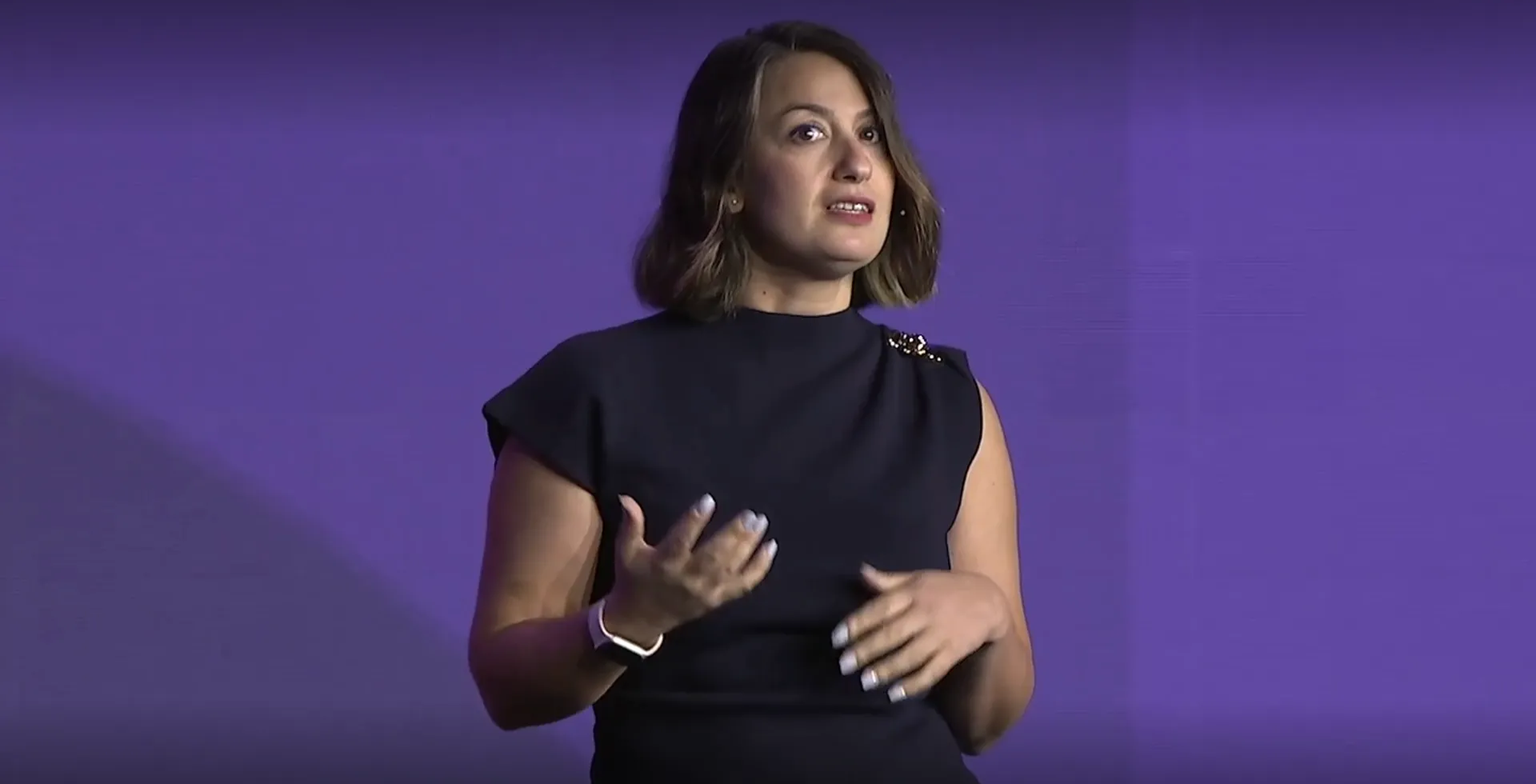 Person (Ezgi Gocucu) speaks on stage in front of a purple background, gesturing with a hand.