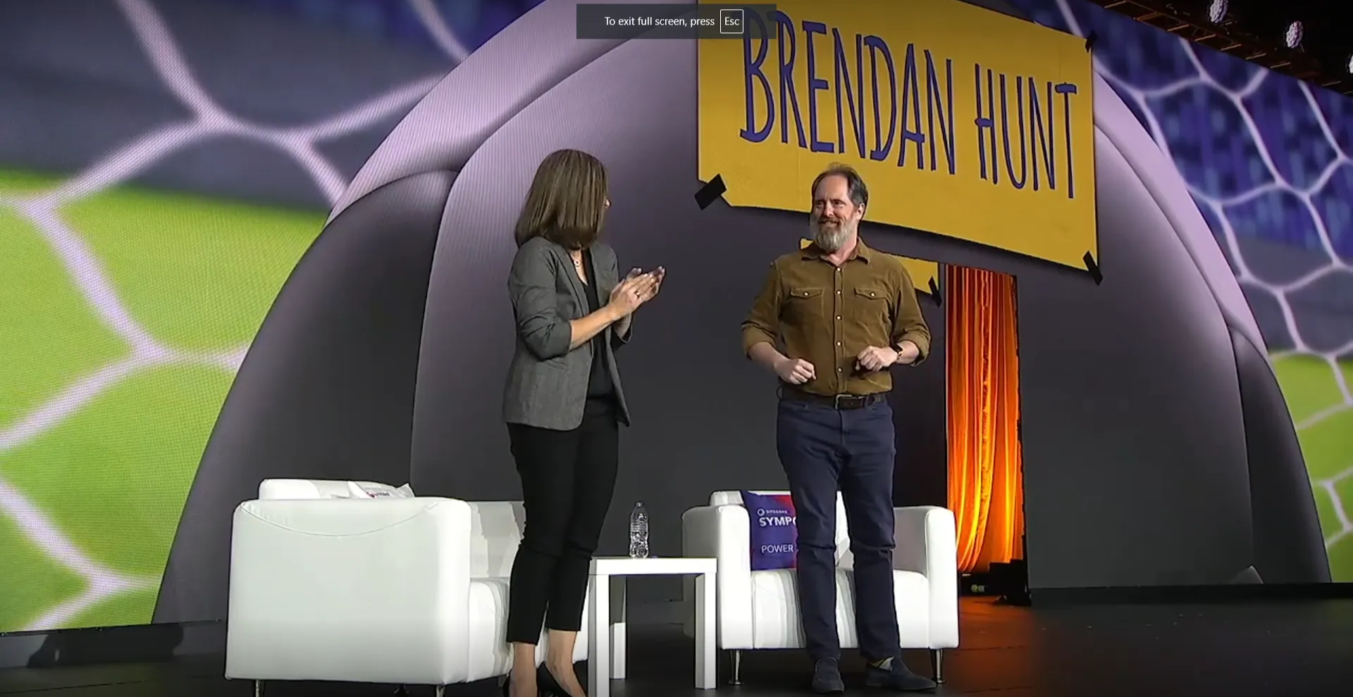 Two people (Hannah Grap and Brendan Hunt) standing on stage in front of visuals that show the name Brendan Hunt. Hannah is clapping for the actor to thank him for his appearance.