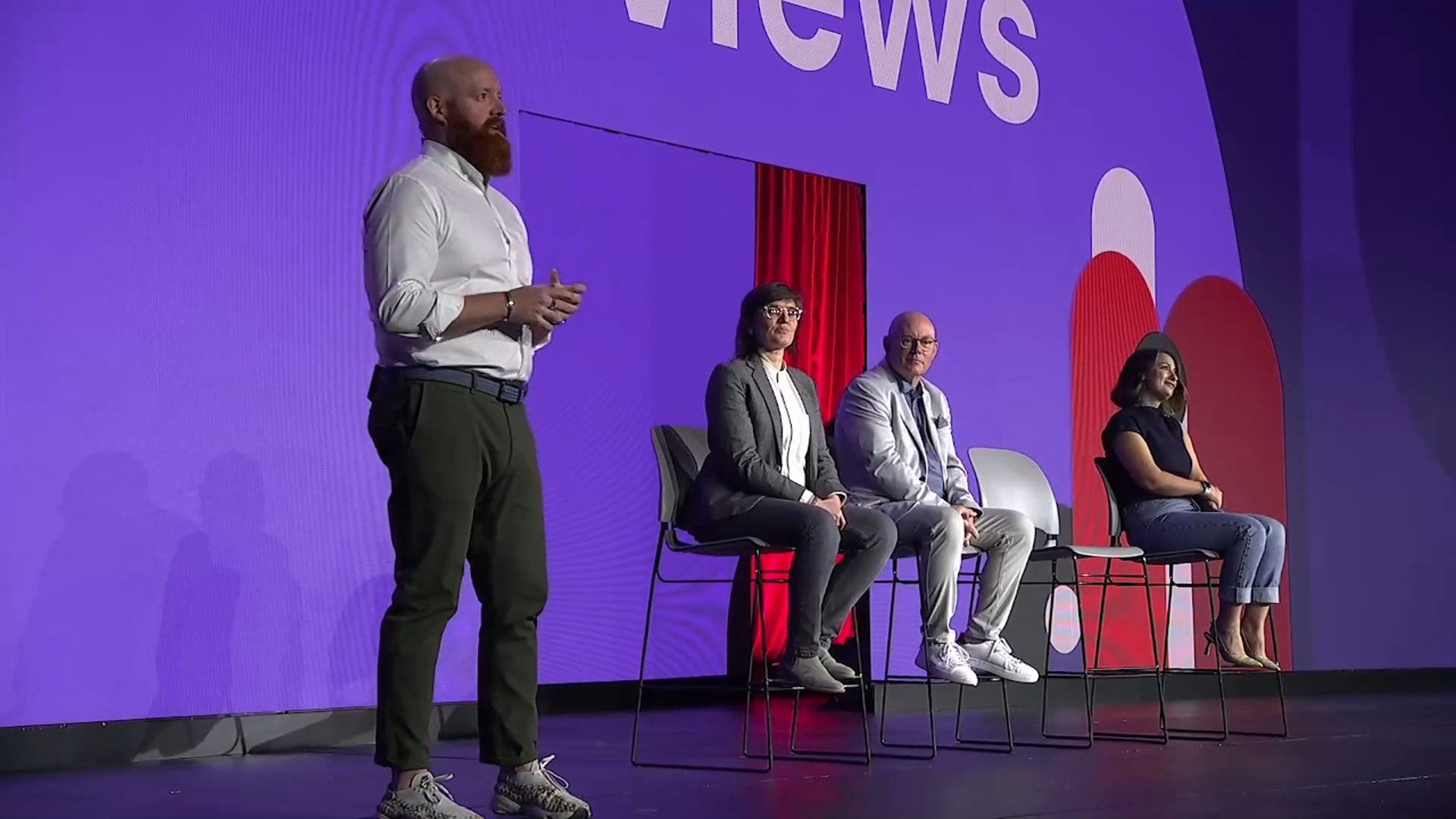 Bearded individual in white shirt, Ru Barry, stands on stage. Three other speakers sit on chairs in the background. Symposium 2024 in Nashville