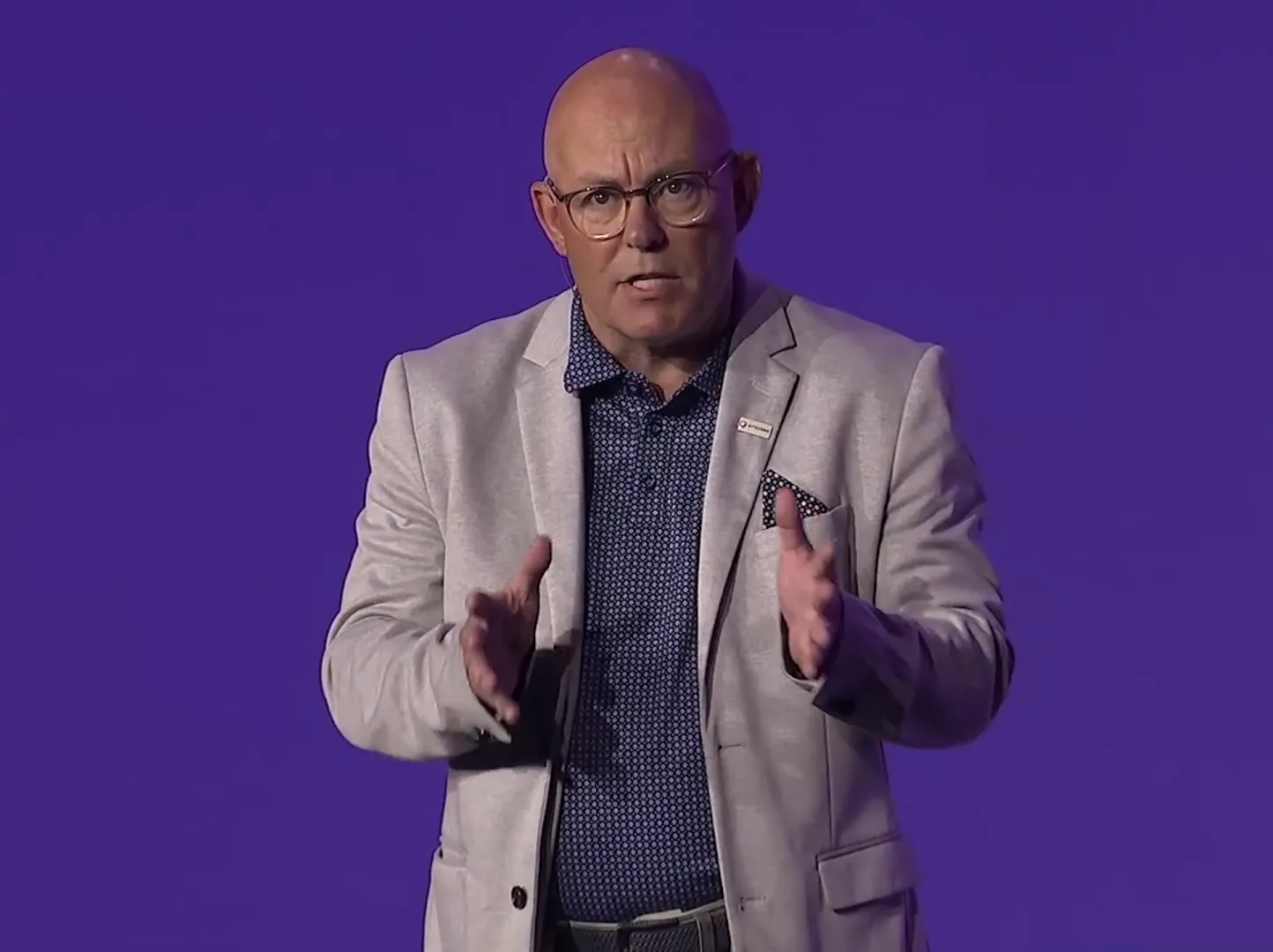 Bald person in glasses, Steve Davis, stands in front of purple background, on stage at Sitecore Symposium 2024 in Nashville