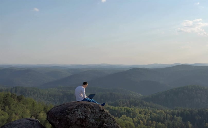 Ein Mann sitzt auf einem Felsen und benutzt einen Laptop, umgeben von Natur
