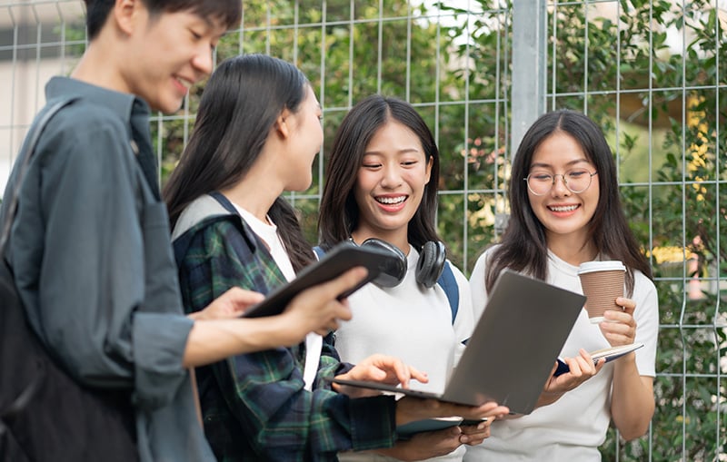 屋外でノートパソコンやタブレットを見ながら笑い合う学生たち