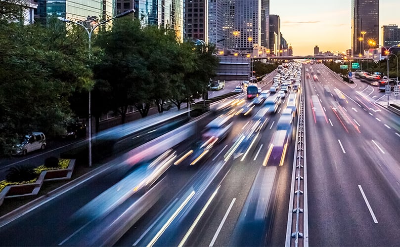 夕暮れ時の都市高速道路を、車のライトが軌跡を描きながら走り抜けている様子。
