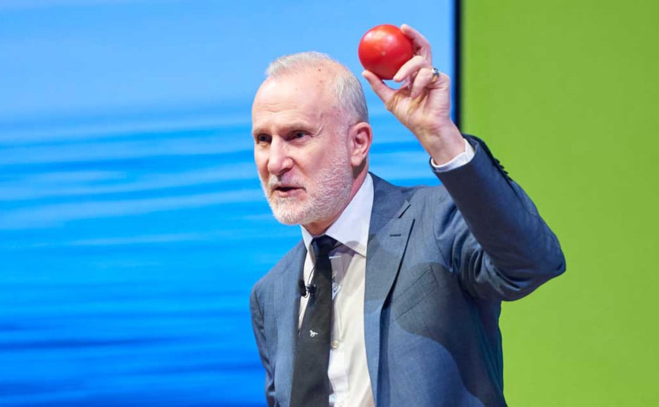 白い髪と髭を生やした、スーツを着た男性(BWT社のCEO、テリー・ポール氏)がトマトを片手に講演を行う様子