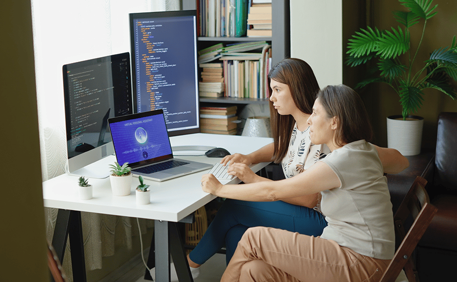 2人の女性が自宅のデスクで複数のモニターを見ながら、コンピューターで作業している。