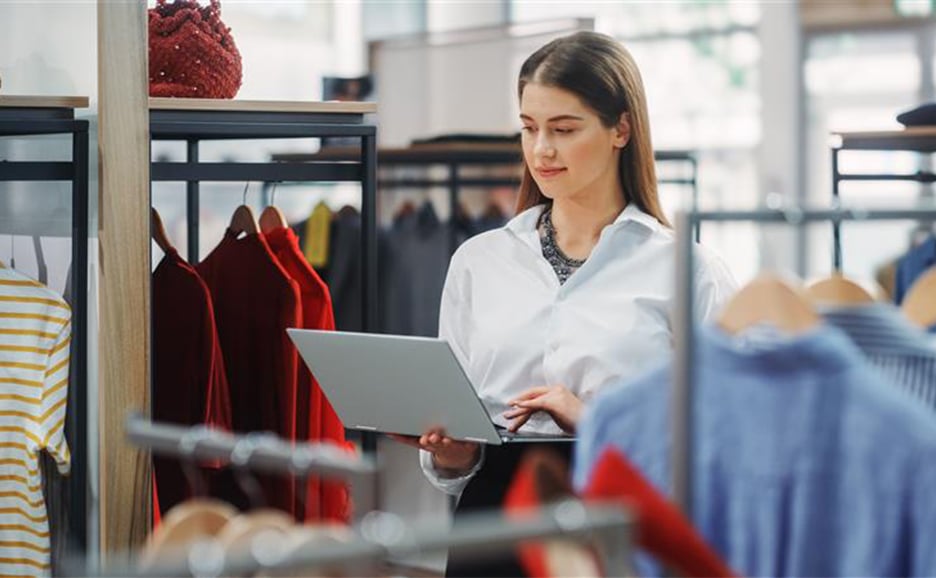 女性が衣料品店の中で、洋服の間に立ち、ノートパソコンを操作しているところ