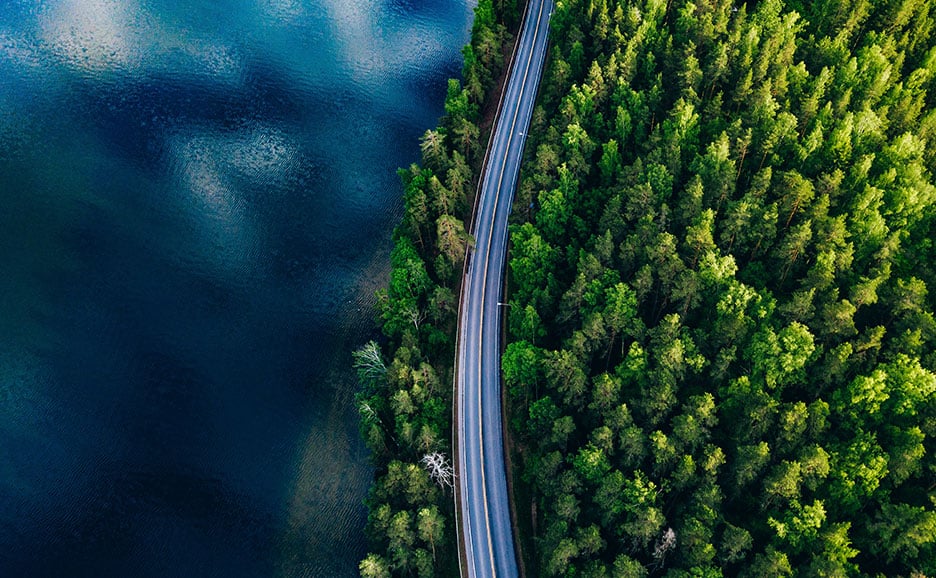 Aerial view of a road stretching between a deep blue lake and a lush green forest.