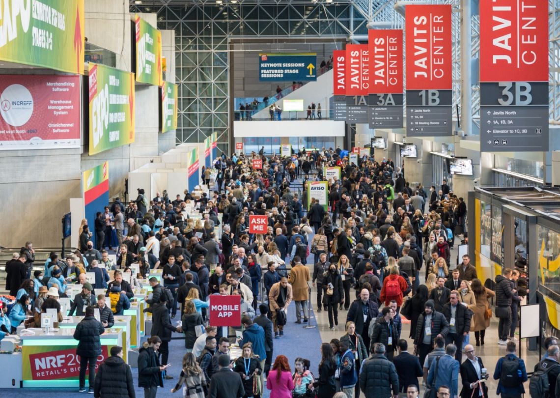Imagem de uma feira lotada no Javits Center, com pessoas interagindo em estandes e banners promocionais ao fundo para a NRF 2023.