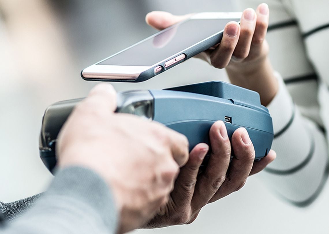 Imagem de pagamento com smartphone utilizando tecnologia de proximidade. Duas mãos segurando um celular e um dispositivo de pagamento.