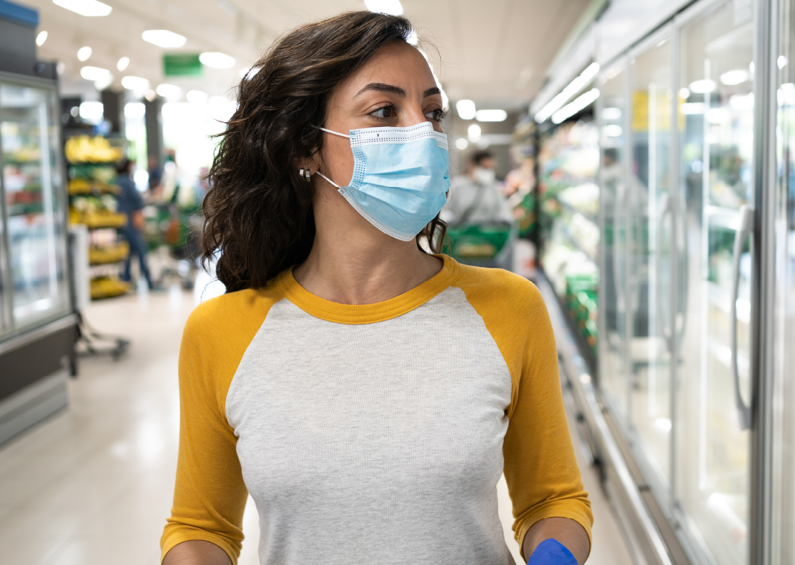 Mulher usando máscara no supermercado, observando opções na prateleira. A imagem retrata a importância da proteção durante as compras.