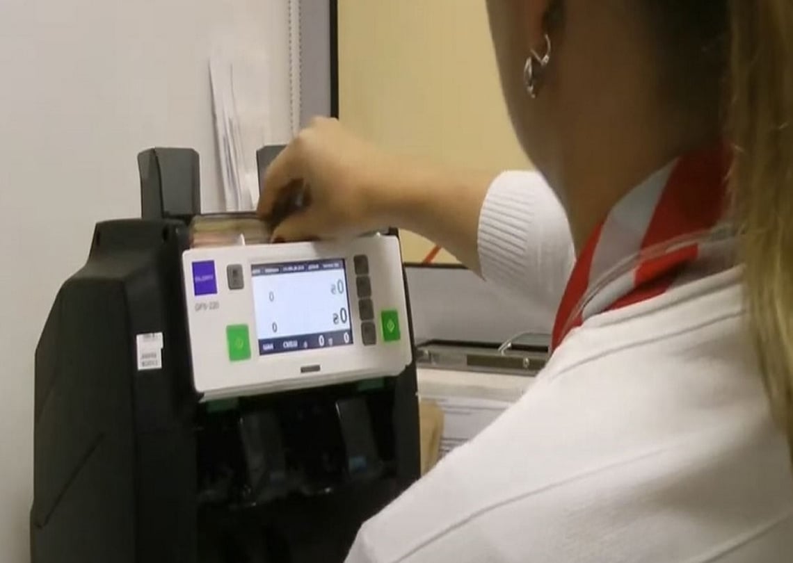 Funcionário usando uma máquina de contagem de dinheiro (GFS-220) para verificar e contabilizar notas em um ambiente bancário.