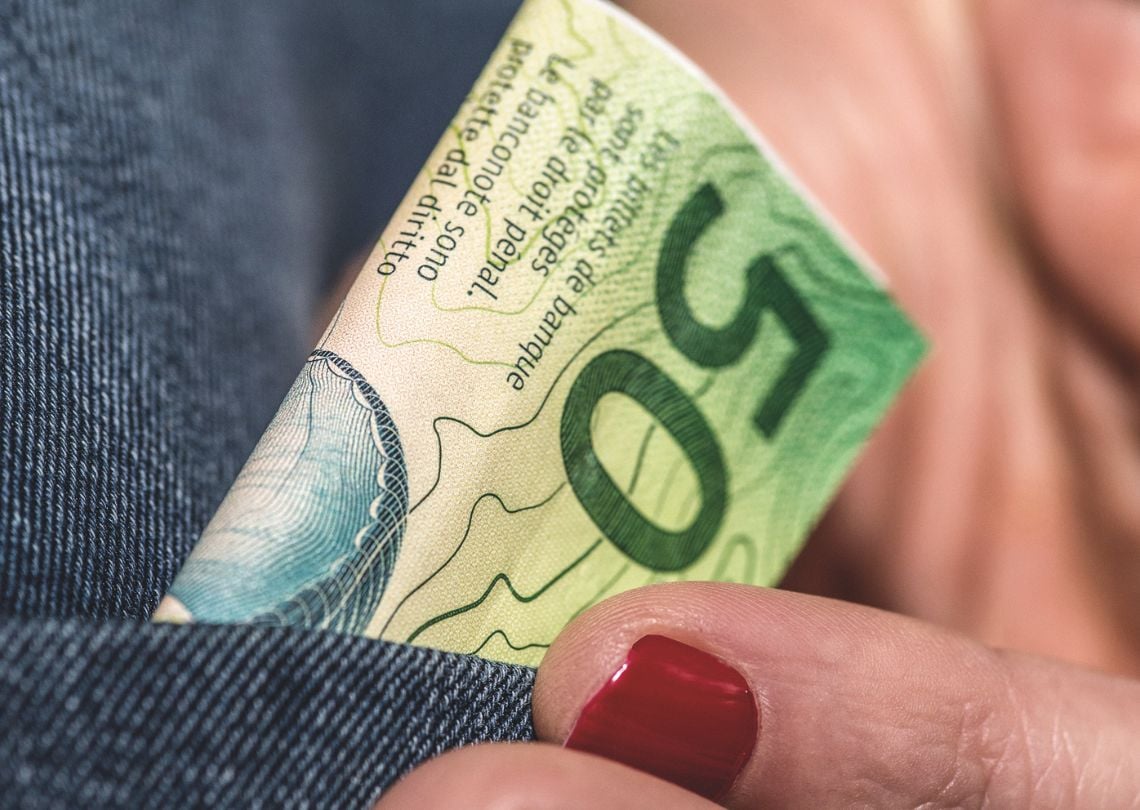Nota de cinquenta sendo retirada do bolso de uma calça jeans. O foco está no detalhe da nota e nas unhas vermelhas da mão que a segura.