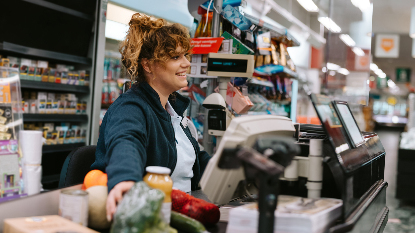 Funcionária de supermercado sorrindo enquanto atende no caixa, rodeada por produtos frescos, demonstrando um excelente atendimento ao cliente.