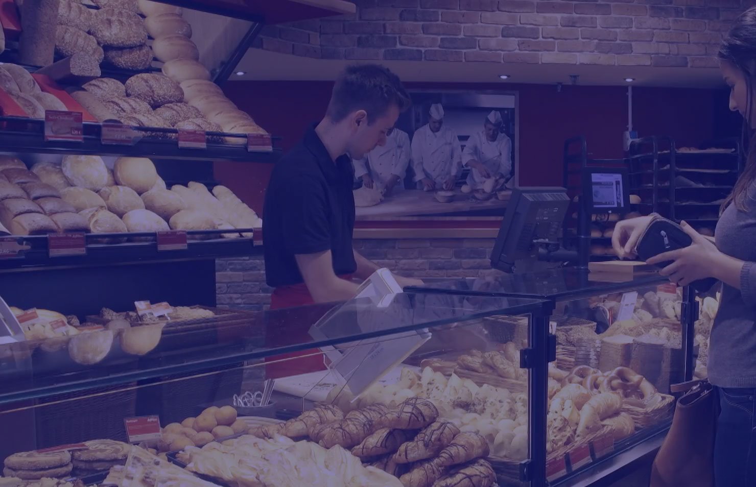 Imagem de uma padaria com diversas opções de pães, onde um atendente serve uma cliente que consulta o menu. Uma variedade de pães é exibida em uma vitrine atraente, destacando a qualidade e frescor dos produtos.