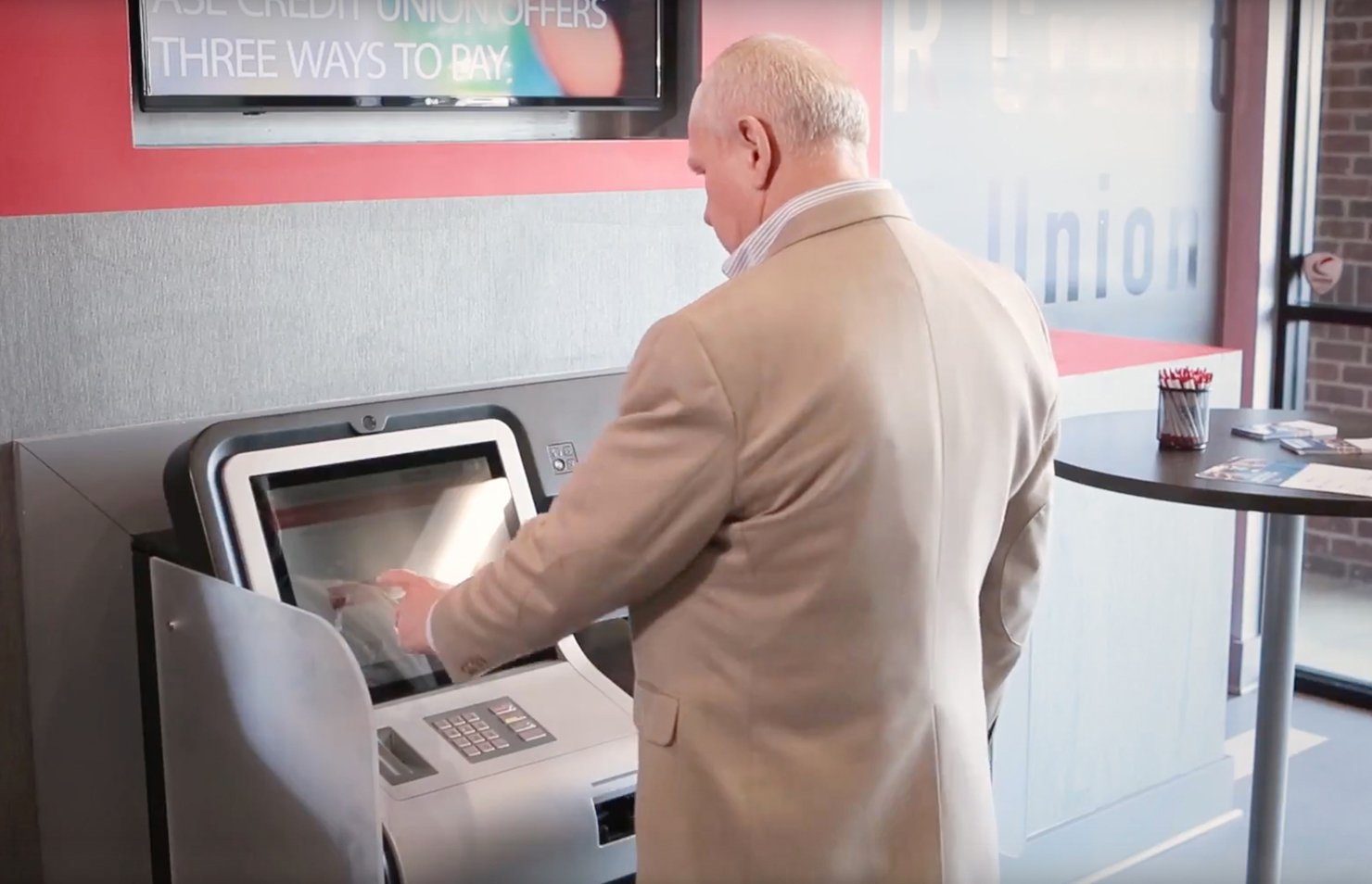 Homem utilizando um caixa eletrônico em uma instituição financeira, interagindo com a tela touch para realizar transações. A imagem mostra um ambiente moderno e acessível.