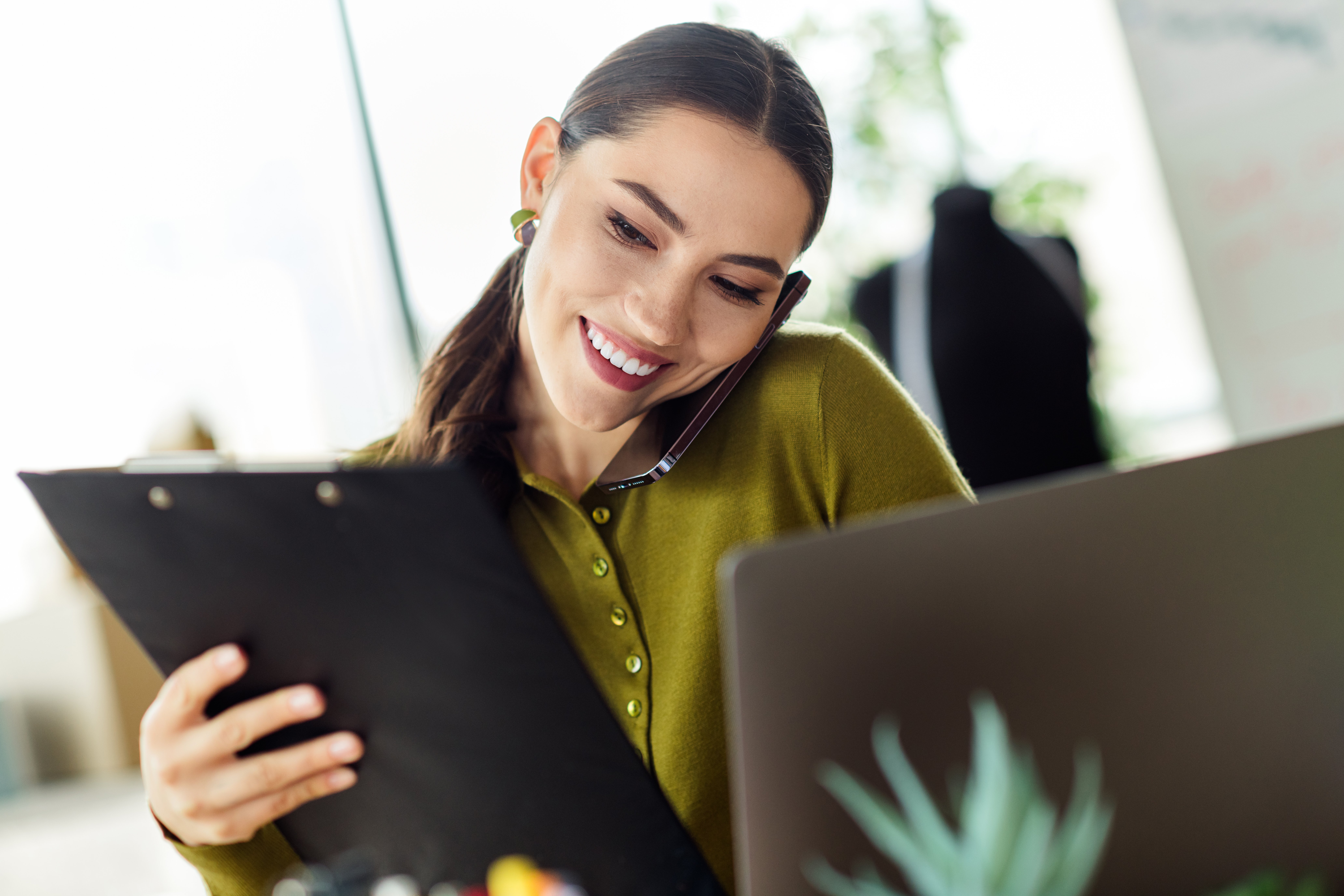 A smiling professional woman multitasks on a phone call while reviewing documents at her desk.