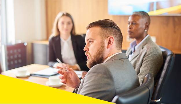 Three professionals seated in a meeting room with laptops and documents while engaging in a discussion.