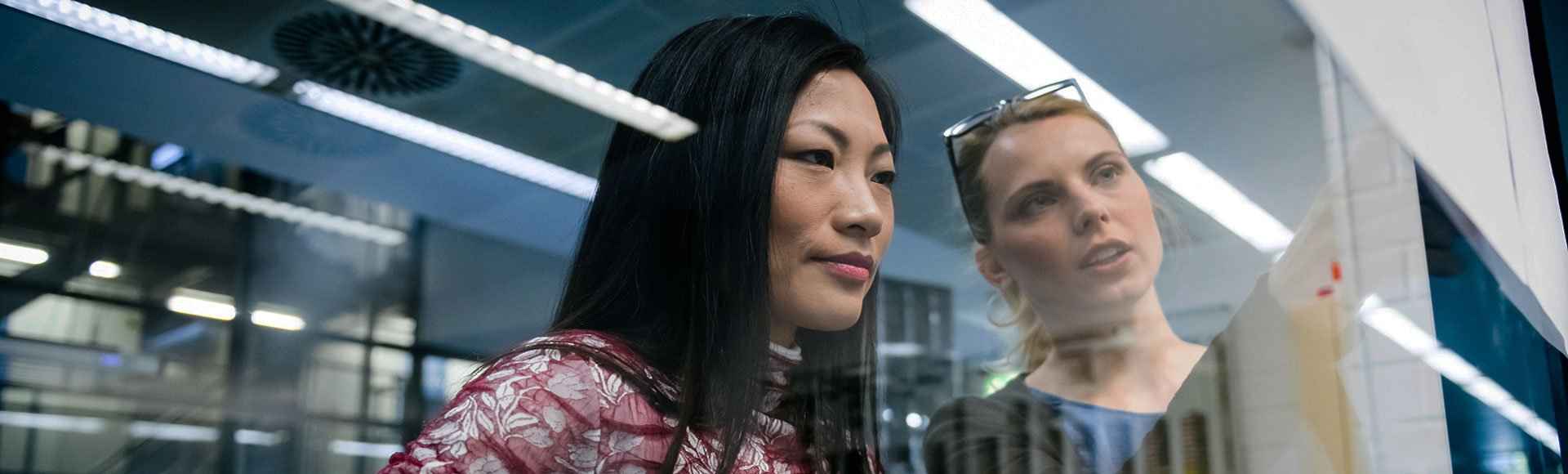 Two female engineer-advisors at Hatch discussing a project in an industrial office while looking at documents pinned on a glass wall.