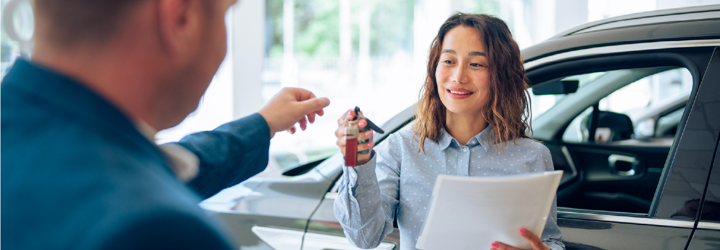 A smiling woman holding documents receives car keys from a person at a dealership, with a dark sedan in the background.