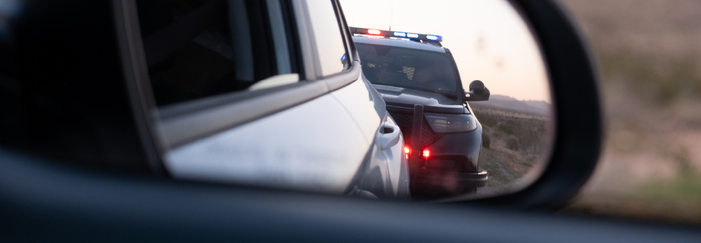 A police car with flashing lights reflected in a vehicle's side mirror on a rural road.