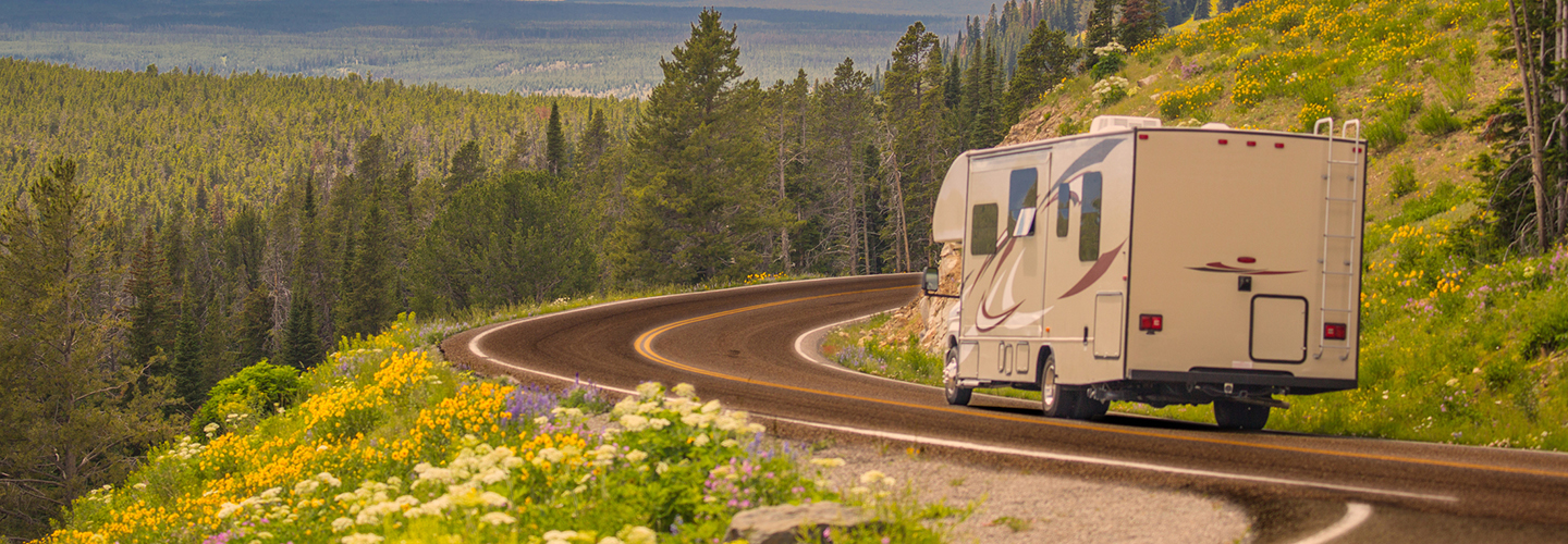 An RV driving along a winding mountain road lined with wildflowers and pine trees under a vibrant sunset sky.