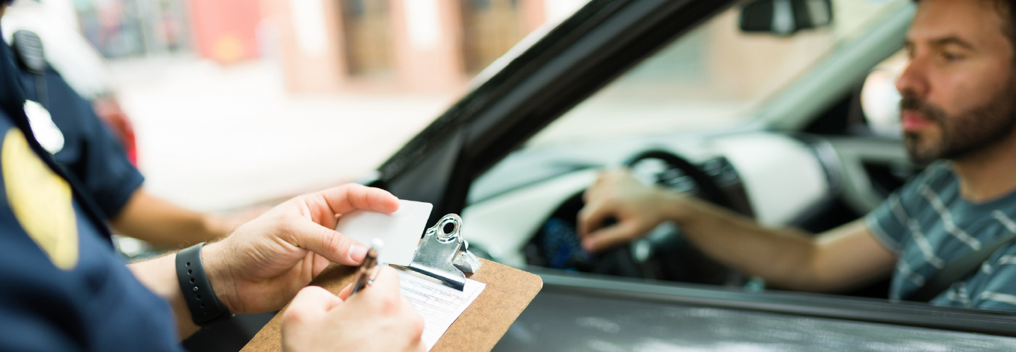 An officer writing on a clipboard while accepting a driver's license from a man seated in a pulled-over vehicle.
