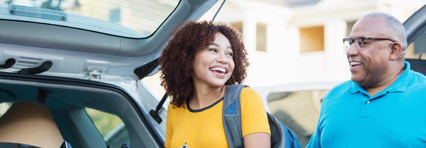A smiling young woman with a backpack and laundry basket unloads a car with an older man, suggesting a student moving away from home.