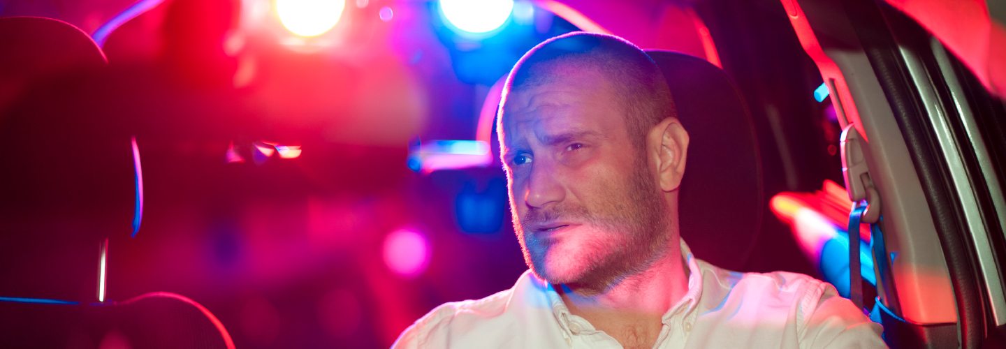A tense man gripping a steering wheel at night, lit by red and blue police lights behind his vehicle.