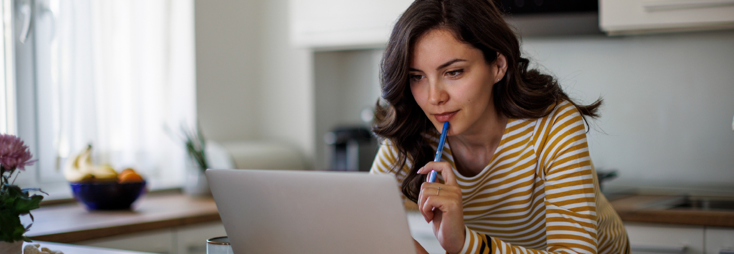 A woman pensively reviews information on a laptop at a kitchen table with a notepad nearby.