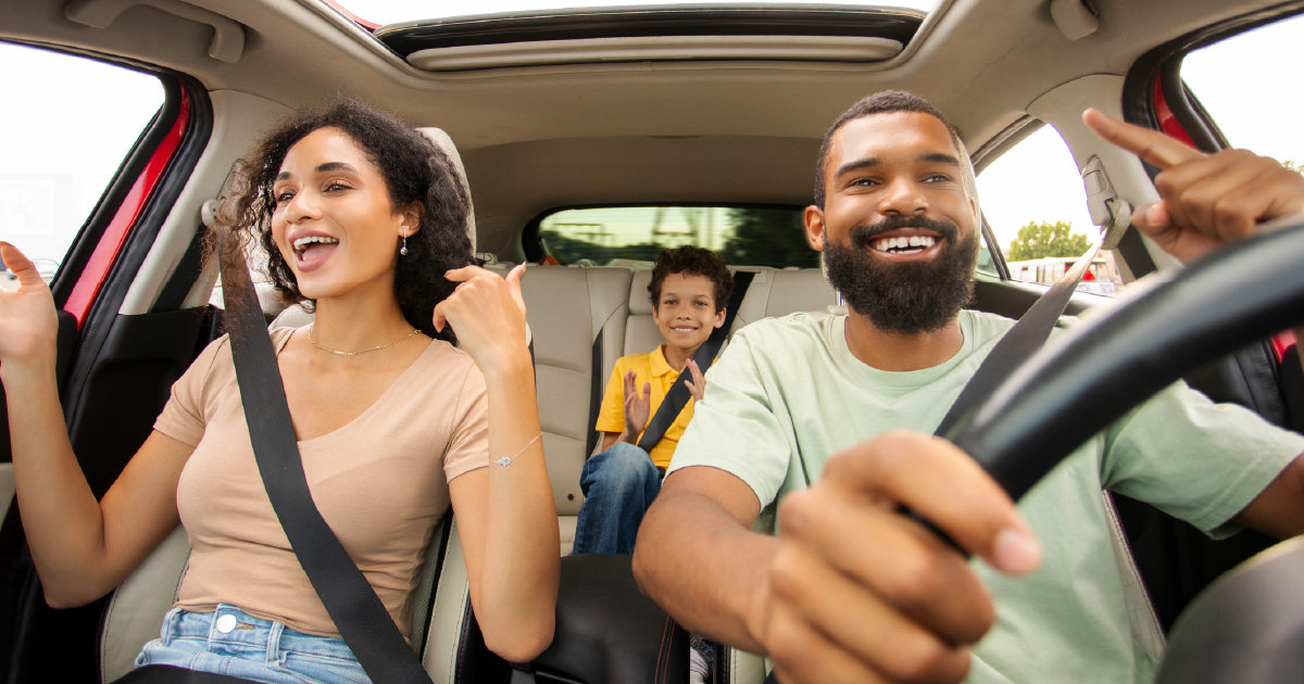 A laughing family of three enjoys a road trip, with two adults in the front seat and a smiling child in the back.