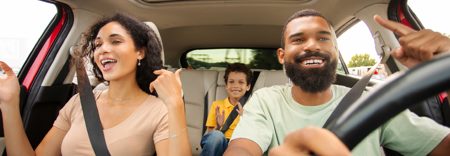A laughing family of three enjoys a road trip, with two adults in the front seat and a smiling child in the back.