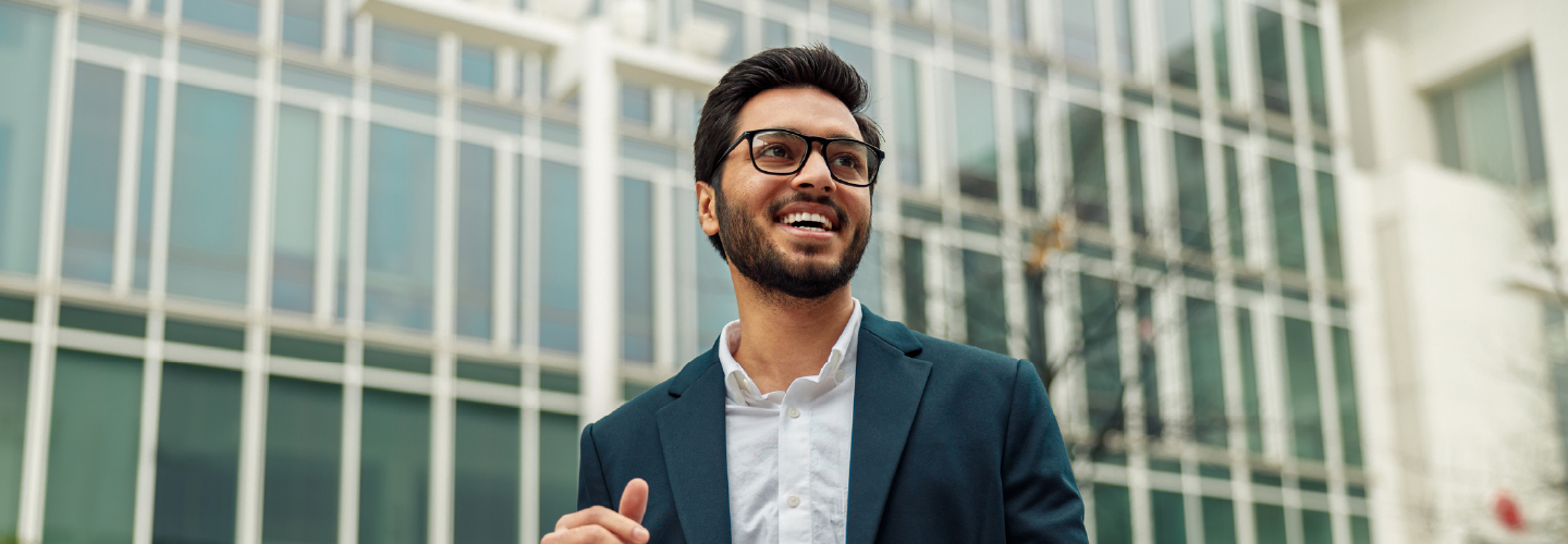A smiling young professional in a blazer holds a laptop outside a modern glass office building.