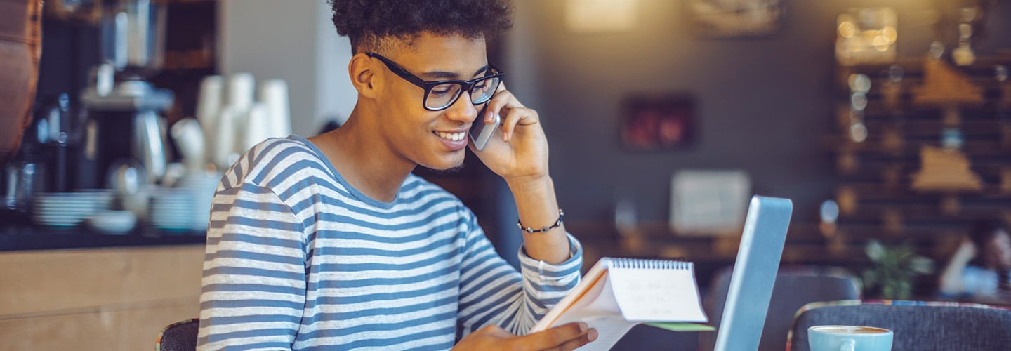 man at a coffee shop on the phone looking at a notebook in his hand with a laptop on the table