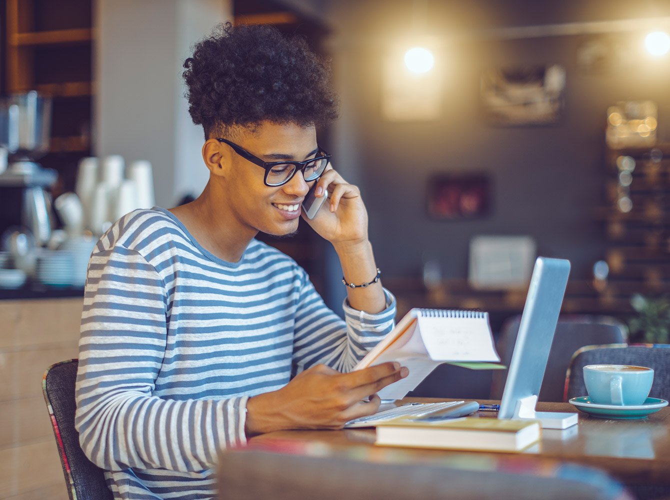 man at a coffee shop on the phone looking at a notebook in his hand with a laptop on the table