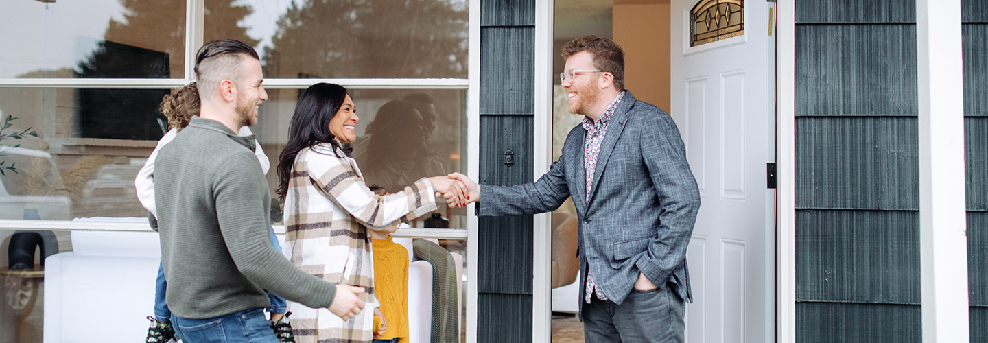 A realtor is shaking hands with a family at the front door of a house.