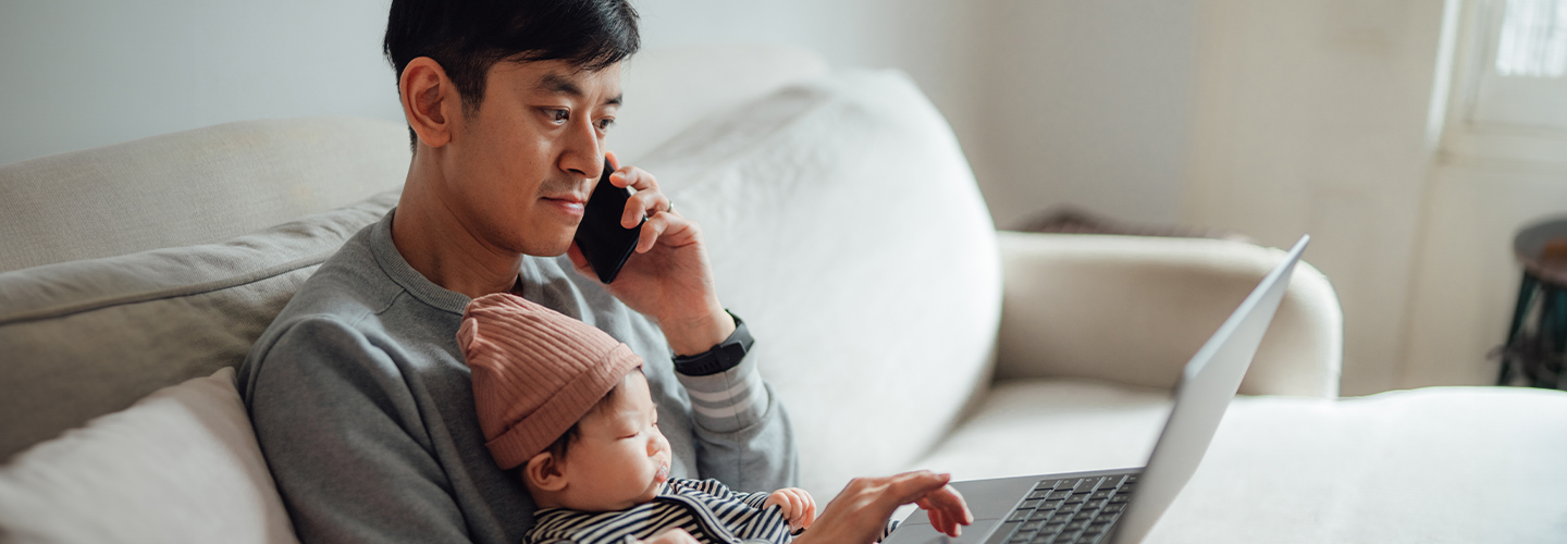 A man with a baby in his lap holds a cellphone to his ear while working on a laptop.